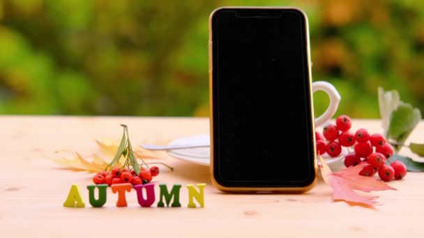 带有空白黑色屏幕的智能手机放在木制桌子上方的茶托上 具有自然绿色背景 户外早茶概念 女性手放置彩色字母词 — 图库视频影像