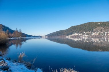 serene winter lakeside scene with snow-covered banks, blue water lake gerardmer, French Vosges, forested hills in distance, frozen landscape snow, still water, and bare trees, Seasonal beauty france