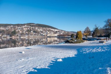 serene winter lakeside scene with snow-covered banks, blue water lake gerardmer, French Vosges, forested hills in distance, frozen landscape snow, still water, and bare trees, Seasonal beauty france