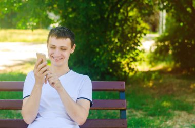 Genç bir Avrupa adam bir şehir parkta bir bankta oturur ve telefon puan bir parmak. Bir adam seyir telefon gülümsüyor. Sevinç ve zevk, neşe ve kahkaha kavramı. Mizah, iletişim.