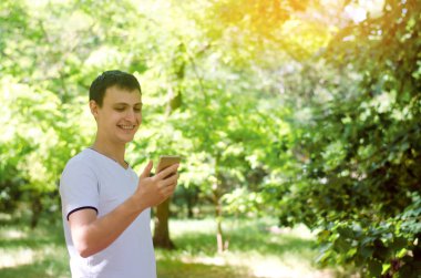 Smartphone bakarak ve gülümseyen genç Avrupa bir adam. telefon bağımlılığı, sosyal ağlar. Internet üzerinde çalışma. mesajı yazın. Doğa, açık. güneşli bir gün. yakın çekim, metin için yer.