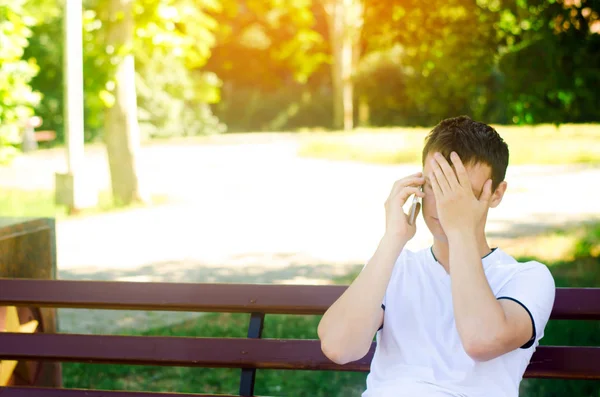 beyaz bir T-shirt genç bir dalgın Avrupa adam telefonda konuşuyor ve şehir parkta bir bankta oturur. yüzünü elleriyle kapsar. adam üzgün, bu beklenmedik Haberler kötü. Sorunları çözme kavramı