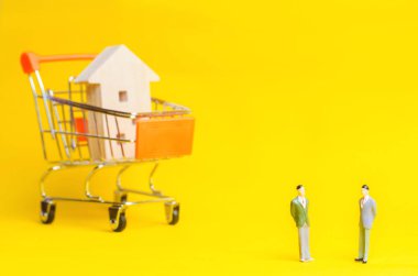 Ahşap bir ev ve bir süpermarket sepetten arka plan üzerinde insanlar stand. Emlak kavramı. Satın alma, satış ve bir ev kiralama. Kredi bir daireye, ipotek oranı için. Genç aileler için uygun fiyatlı konut. bir Emlak Hizmetleri