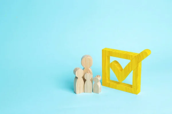 Three wooden human figures stand together next to a tick in the box. The concept of elections and social technologies. Volunteers, parties, candidates, constituency electorates. Human rights.