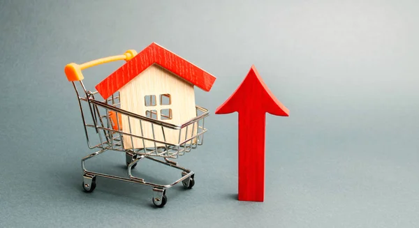 Drewniany dom w wózku supermarket i czerwona strzałka w górę. Koncepcja zwiększania kosztów mieszkaniowych. Wysokie zapotrzebowanie na nieruchomości. Wzrost czynszu i oprocentowanie kredytów hipotecznych. Sprzedaż mieszkań — Zdjęcie stockowe