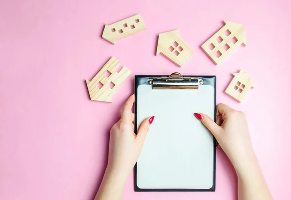 The girl holds an empty tablet for records on a pink background with many wooden houses. A real estate agent is looking for or offering housing options for a client. Search for a house or apartment.