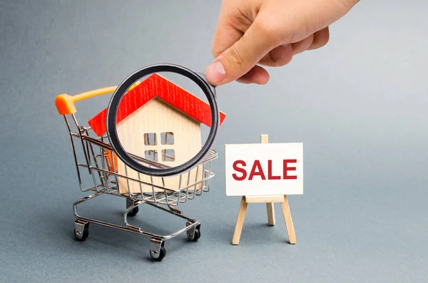 Förstoringsglaset är att titta på snabbköpet vagn med hus och en försäljning affisch. Konceptet att sälja ett hem, fastigheter tjänster eller köpa från ägaren. fastighet, lägenheter. Selektivt fokus — Stockfoto
