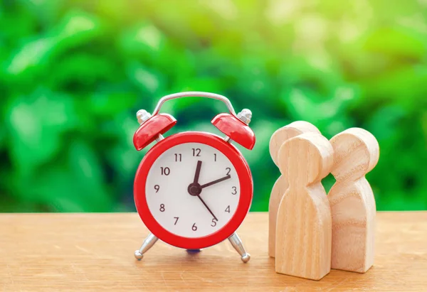 Tres figuras humanas de madera de pie junto a un despertador rojo sobre un fondo natural. Concepto de tiempo. Negocios, pago por hora. Horas de hombre. Planeando. minimalismo — Foto de Stock