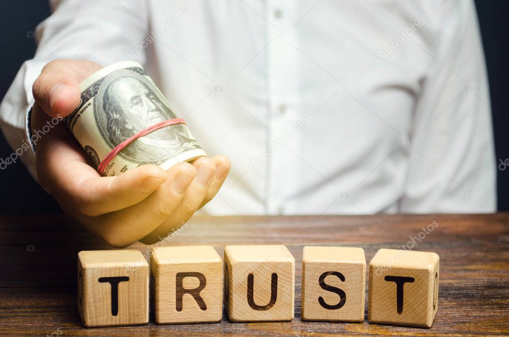 Wooden blocks with the word Trust and money in the hands of a businessman. Trust relationships between business partners, friends, relatives. Respect and authority. Confidence in a person.