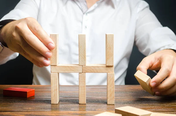 L'uomo d'affari colleziona una casa di domino. Creazione concettuale di una nuova impresa o di una nuova impresa, instaurazione di processi. Costruzione di un edificio residenziale. Ipoteca. Mercato immobiliare e investimenti . — Foto Stock