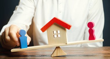 Terazide bir erkek ve bir kadının ahşap figürleri ve aralarında bir ev. Yasal yollarla mal paylaşımı. Konutun mülkiyetinin açıklanması. Boşanma kavramı. Avukatım. Mahkeme. Eşler