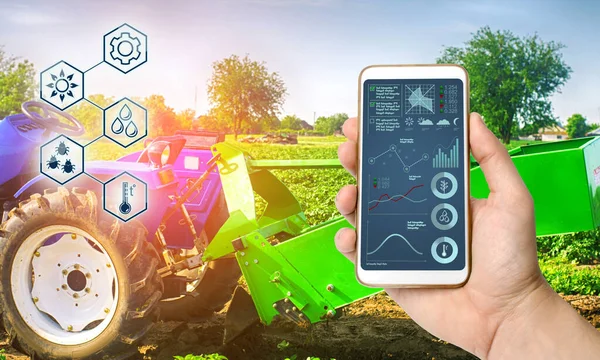 Farmer holds smartphone with infographic on tractor background with potato digger. Farming and smart agriculture. Agricultural machinery, data analyzing on plants status. Harvesting.