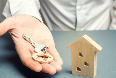 Bir adamın elinde evi ve ahşap evi olan bir ıvır zıvır anahtarı var. Emlak konsepti. Emlakçı servisi. Mülk satışı. Ev al. Kiralık daireler. İpotek. Satılık ev. İnşaat yapılıyor.