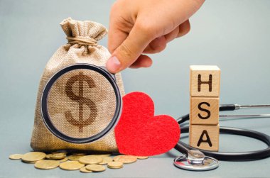 HSA sözcüğüyle tahta bloklar ve steteskoplu para çantası. Sağlık tasarruf hesabı. Sağlık hizmetleri. Sağlık sigortası. Yatırımlar. Vergisiz tıbbi harcamalar. Bozuk para ve dolar işareti. Kırmızı kalp