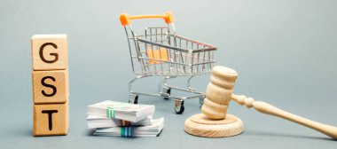 GST sözcüğüyle tahta bloklar, para ve yargıç tokmağı olan bir market arabası. Vergi, mal ve hizmetlerin satışına dayatılıyor.