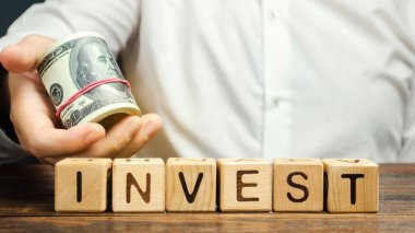 Yatırım sözcüğüyle tahta bloklar ve bir işadamının elinde para. Bir iş projesine yatırım yapma fikri. Ekonomi ve finans. Kar için nakit yatırımlar