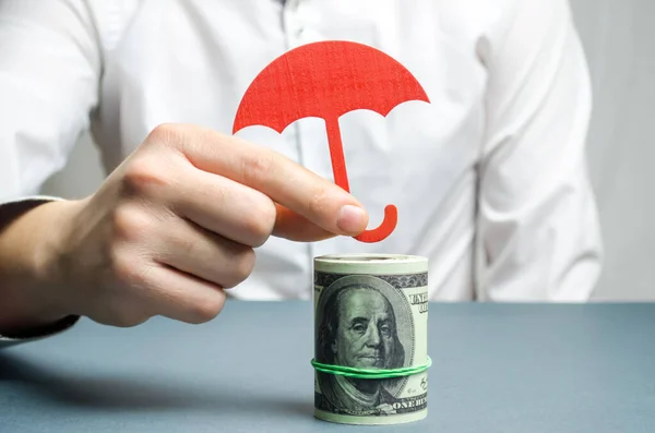 An insurance agent holds a red umbrella over dollar bills. Savings protection. Keeping money safe. Investment and capital insurance. The risk of doing business. Corruption losses. Arrest of investors