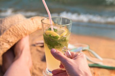 Kafkas kadın sahil kumlu plaj gün batımında oturur ve mojito kokteyl elinde tutar. Bir sandal ve bir arka plan, şapkası