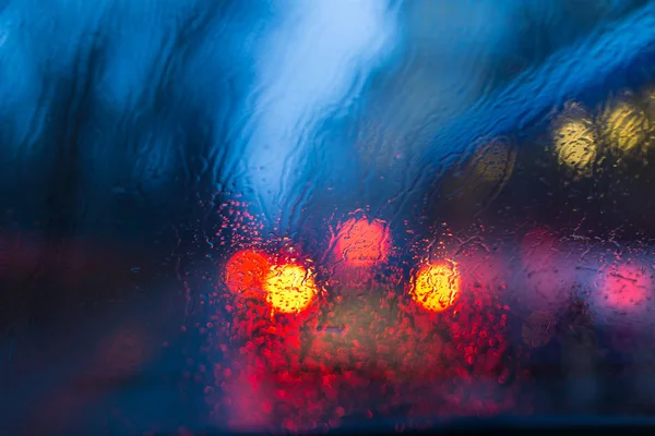 Bokeh aydınlatma cam araç ve bir yağmur ile gece şehrin yangınların üzerinden. Bir yağmur akışını camın üzerine düşer. Belirsiz odak