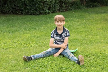 Kot pantolon ve mavi çizgili tişört giyen beyaz bir çocuk parktaki yeşil çimlerin üzerinde oturuyor..
