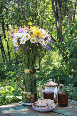 Durgun bir hayat, masada kır çiçekleriyle dolu bir vazo, bir fincan çay ve dilimlenmiş kek var.