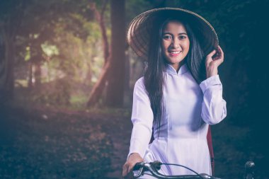 Bir bisiklet ve bir şapka tarafından yemyeşil bahçeleri arka plan smog ortasında ve güneş sabah holding halen Vietnam (Ao dai) giyinen uzun saçlı güzel genç Asyalı teen portresi çekili.