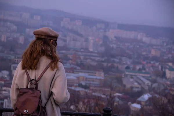 Una chica solitaria con una mochila de cuero en una gorra está de pie en una alta montaña y mirando la ciudad . — Foto de Stock