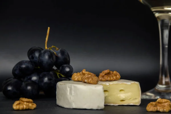 Close-up chopped camembert cheese, nuts and sweet blue grapes on the background of a glass of white dry wine — Stock Photo, Image