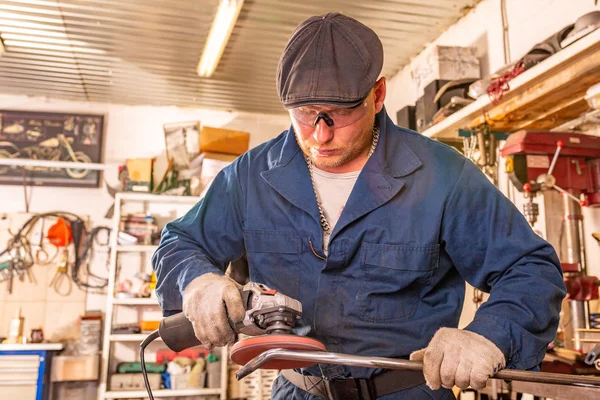 Bir genç adam kaynakçı mavi tişört, gözlük ve inşaat eldiven süreçleri metal içinde belgili tanımlık geçmiş birçok araç garajda bir açı değirmeni — Stok fotoğraf