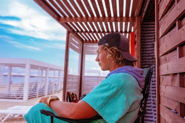 Joven surfista chico atractivo con un albornoz está sentado en la terraza junto al mar, relajante y mirando a la distancia en el mar —  Fotos de Stock