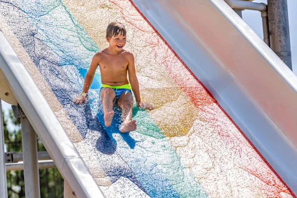 Um menino feliz no slide de água em uma piscina se divertindo durante as férias de verão em um belo parque aquático. Um rapaz a escorregar pelo escorrega de água e a fazer salpicos. — Fotografia de Stock