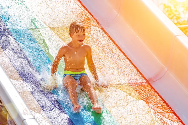 Egy boldog fiú csúszdán egy medencében szórakozik a nyári vakáció alatt egy gyönyörű aquaparkban. Egy fiú lecsúszott a csúszdán és fröcskölt.. — Stock Fotó