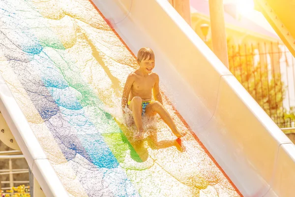 Um menino feliz no slide de água em uma piscina se divertindo durante as férias de verão em um belo parque aquático. Um rapaz a escorregar pelo escorrega de água e a fazer salpicos. — Fotografia de Stock