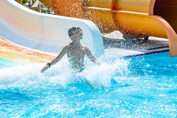 아름다운 아쿠아 공원에서 여름 방학을 즐기며 수영장에서 워터 슬라이드를 타고 있는 행복 한 소년. 한 소년이 물을 미끄러져 내려오면서 물줄기를 만들고 있었다. — 스톡 사진