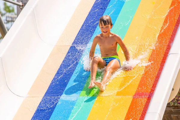 Um menino feliz no slide de água em uma piscina se divertindo durante as férias de verão em um belo parque aquático. Um rapaz a escorregar pelo escorrega de água e a fazer salpicos. — Fotografia de Stock