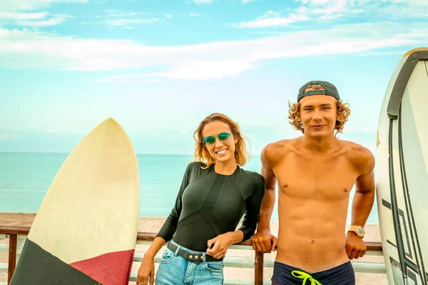 Χαμογελώντας νεαρό ζευγάρι ενεργό surfers χαλαρώνοντας στην παραλία μετά τον αθλητισμό με σανίδα του σερφ. Υγιεινού τρόπου ζωής. Ακραία σπορ — Φωτογραφία Αρχείου