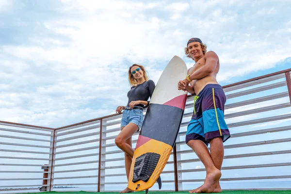 Χαμογελώντας νεαρό ζευγάρι ενεργό surfers χαλαρώνοντας στην παραλία μετά τον αθλητισμό με σανίδα του σερφ. Υγιεινού τρόπου ζωής. Ακραία σπορ — Φωτογραφία Αρχείου