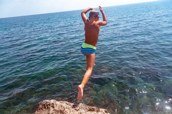 Un niño está saltando desde el acantilado hacia el mar en un caluroso día de verano. Vacaciones en la playa. El concepto de turismo activo y recreación —  Fotos de Stock