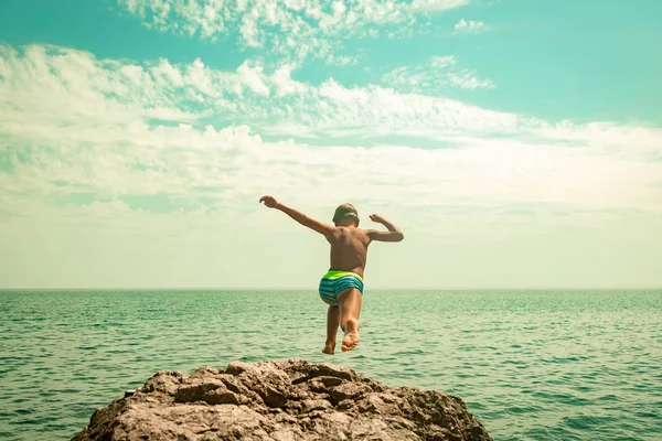 Un niño está saltando desde el acantilado hacia el mar en un caluroso día de verano. Vacaciones en la playa. El concepto de turismo activo y recreación —  Fotos de Stock