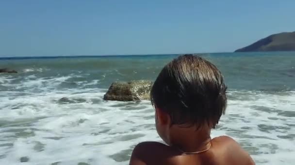 Een jongen zit aan het strand in het zand en kijkt naar het water in en uit komen — Stockvideo