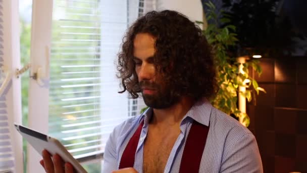 Чоловік читає новини на планшетному комп'ютері і п'є каву на кухні — стокове відео