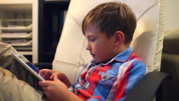 Щасливий молодий хлопчик грає на планшеті, сидячи на стільці вдома . — стокове відео