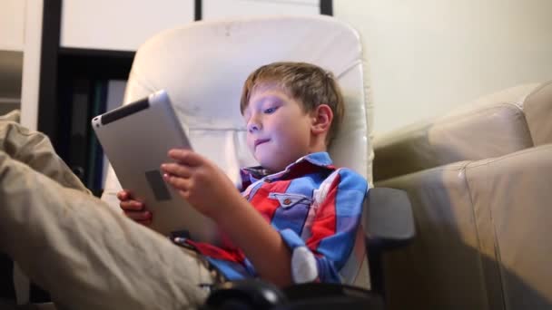 Щасливий молодий хлопчик грає на планшеті, сидячи на стільці вдома . — стокове відео