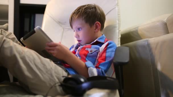 Gelukkige jongen speelt op zijn tablet zitten op de stoel thuis. — Stockvideo