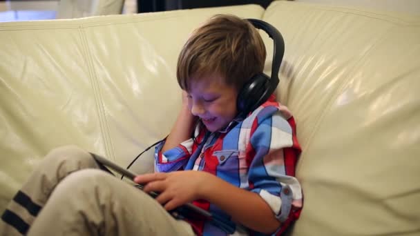 Niño feliz en los auriculares juega en la tableta y escucha música mientras está sentado en el sofá en casa — Vídeos de Stock