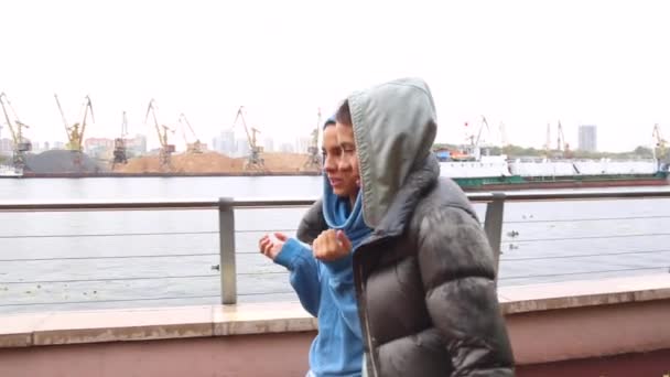 Duas meninas bonitas gêmeas em sportswear estão andando ao longo do dique do rio na chuva, escondendo-se atrás de uma jaqueta, sentindo-se feliz. Jovens mulheres vestindo pano quente, apreciando a natureza — Vídeo de Stock