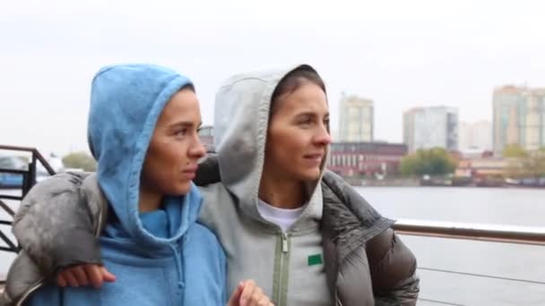 Две красивые девушки-близнецы в спортивной одежде идут по набережной реки под дождем, прячась за одной курткой, чувствуя себя счастливыми. Молодые женщины в теплой одежде, наслаждаясь природой — стоковое видео