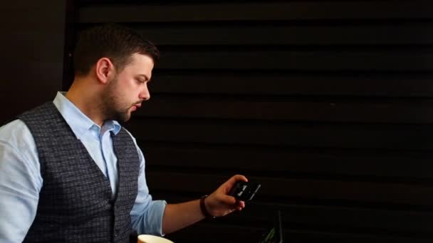 Фрилансер-бизнесмен работает на ноутбуке с диаграммами на экране в кафе, проверяет приложение смартфона на наличие информации и обновлений, а также пьет кофе. Отслеживание и выявление выстрела — стоковое видео