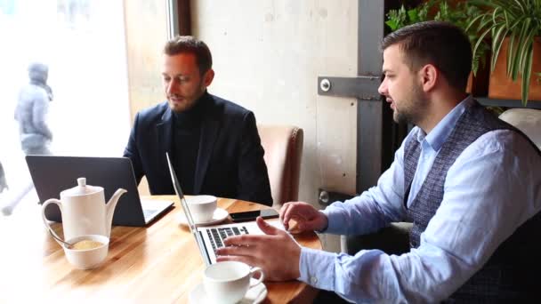 Два бизнесмена сидят за столиком кафе, пользуются ноутбуком, пьют чай и разговаривают — стоковое видео