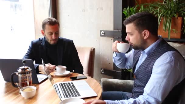 Dos hombres de negocios sentados en la mesa de la cafetería, usando el ordenador portátil, tomando té y hablando — Vídeos de Stock